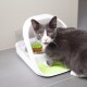 Eine graue Katze mit gelben Augen sitzt neben einem modernen Haustierfutterspender mit einer grünen Schüssel, die mit Trockenfutter gefüllt ist.