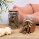Zwei Glasbehälter gefüllt mit Hundeleckerlis, einer umgekippt und verschüttet Leckerlis, neben einem beigen Seilspielzeug, mit einem rosa Haustierbett im Hintergrund.