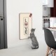 Eine graue Katze steht auf ihren Hinterbeinen und schaut neugierig auf ein wandmontiertes Kratzbrett mit "LIEBE" und einer Katzensilhouette.