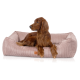 Langhaariger brauner Hund, der entspannt in einem roséfarbenen Cord‑Hundebett liegt, die Vorderpfoten über den Rand gelegt, mit kleinem braunem Etikett.