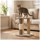 Getigerte Katze steht auf einem beigen runden Kratzbaum mit hängenden Bommelspielzeugen in einem sonnendurchfluteten Wohnzimmer neben einem Sofa und einer Pflanze.