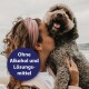 Eine Frau mit lockigem Haar hält einen flauschigen Hund vor einem bewölkten Strandhintergrund, mit einem kreisförmigen Textüberlagerung, der besagt 'Ohne Alkohol und Lösungsmittel.'