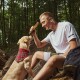 Eine Person in einem weißen T-Shirt und Shorts sitzt auf einem Baumstamm im Wald und hält einen Kaustick in Richtung eines Golden Retrievers, der ein rotes kariertes Halstuch trägt.