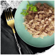 Une assiette teal contient de la nourriture humide pour animaux avec de la viande hachée dans un bouillon, garnie de coriandre, à côté d'une fourchette dorée sur un tissu sombre.