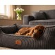 Brauner Hund schläft in grauem Cord-Haustierbett mit Lederanhänger 'KNUFFELWUFF' in einem Wohnzimmer.