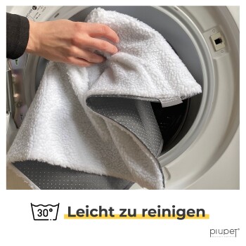 Eine Hand legt eine flauschige weiße Haustierdecke mit einer grauen gepunkteten Unterseite in eine Waschmaschine. Text: "30° Einfach zu reinigen."