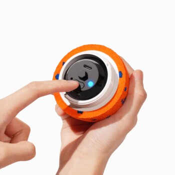 Hände halten einen orangefarbenen Schaumstoff-Haustierball mit schwarzem Elektronikkern, ein Finger drückt einen blau leuchtenden LED-Knopf.