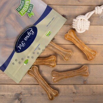 Eine Tüte Hundeleckerlis mit knochenförmigen Kaustücken, die auf einer Holzoberfläche verstreut sind, mit einem Seilspielzeug in der Ecke.