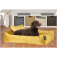 Ein glücklicher brauner Labrador-Retriever, der auf einem weichen gelben Hundebett mit zwei passenden Kissen entspannt, vor einem hellen Raum.