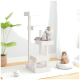 Ein weißer Kratzbaum mit drei Plattformen; eine flauschige graue Katze sitzt oben, und eine orangefarbene Katze entspannt sich auf einem Kissen in der Nähe. Eine grüne Pflanze steht dahinter.