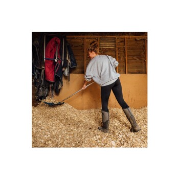 Eine Person in einem grauen Sweatshirt und schwarzen Leggings schaufelt Holzspäne in einem Stall und trägt schlammige Gummistiefel.