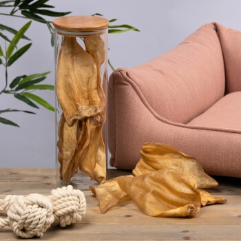 Ein Glasbehälter voller getrockneter Tierkausnacks steht auf einem Holztisch neben einem rosa Haustiersofa und einem weißen Seilspielzeug.
