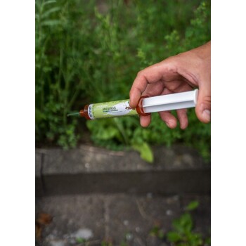 Eine Hand hält ein grünes Röhrchen mit der Aufschrift "AMEISENGEL" mit einer Düse, umgeben von Grün, was auf ein Haustierpflegeprodukt hindeutet.