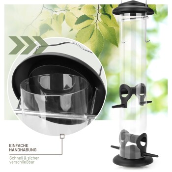 Transparente Vogelhausfütterer mit schwarzem Boden und Oberteil, mit einfacher Handhabung und sicherem Verschluss, umgeben von grünen Blättern.