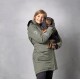 Eine Person in einer grünen Jacke hält einen kleinen, langhaarigen Hund nah an ihrer Brust, vor einem grauen Hintergrund.