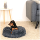 Ein kleiner schwarz-brauner Hund entspannt sich in einem flauschigen grauen Haustierbett auf einem hellen Teppich, mit einer grauen Couch und Lampe im Hintergrund.