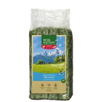 Un sac en plastique transparent rempli de foin vert, étiqueté 'Real Nature' avec un paysage de montagne pittoresque en arrière-plan.
