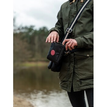 Eine Person in einer grünen Jacke steht an einem Gewässer und greift in eine schwarze Leckerlitasche für Haustiere mit rotem Logo.