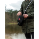 Eine Person in einer grünen Jacke steht an einem Gewässer und greift in eine schwarze Leckerlitasche für Haustiere mit rotem Logo.