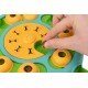 A hand holds a small treat above a colorful interactive pet puzzle toy, featuring yellow and green sections with bone-shaped openings.