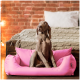 Ein grauer Weimaraner-Hund, der aufrecht in einem hellrosa Haustierbett sitzt, mit einer gemütlichen gestrickten Decke und einem Bett im Hintergrund.