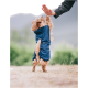 Ein kleiner Hund in einem blauen Outfit steht auf seinen Hinterbeinen und greift nach der Hand einer Person in einer natürlichen Umgebung.