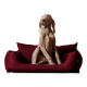 Ein hellbrauner Hund sitzt aufrecht auf einem plüschigen burgunderfarbenen Haustierbett und schaut neugierig zur Seite.