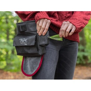 Eine Person in einem roten Pullover hält einen grünen Abfallbeutel, während sie eine schwarze Leckerlitasche mit der Aufschrift 'TruPet TwinTreat Plus' benutzt.