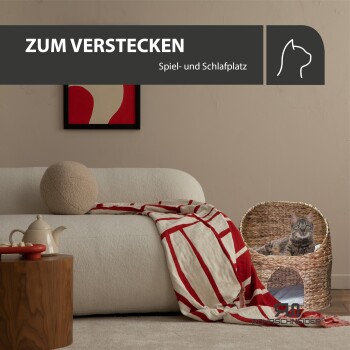 Gemütliches Wohnzimmer mit einem weißen Sofa, einer Katze, die aus einem geflochtenen Bett schaut, einem Holztisch mit Snacks und dem Text "ZUM VERSTECKEN."