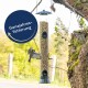 Ein Vogelfutterspender, gefüllt mit Samen, hängt von einem Baum, mit einem kleinen Vogel in der Nähe im Flug. Der Text lautet "Ganzjahresfütterung."