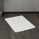 Eine weiße Haustiermatte mit einer strukturierten Oberfläche, die auf dunklem Holzboden neben einer Topfpflanze vor einer grauen Wand liegt.