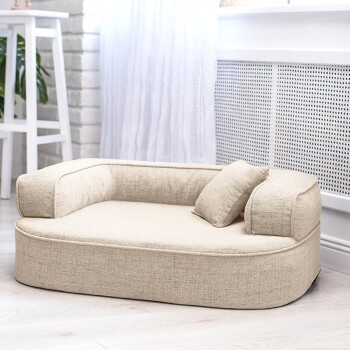 Ein gemütliches beiges Haustierbett mit einem kleinen Kissen, das in einem hellen Raum mit weißen Vorhängen und einer Topfpflanze im Hintergrund platziert ist.