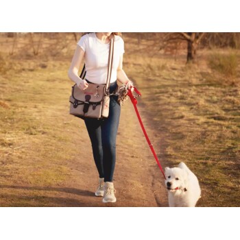 Eine Frau in einem weißen T-Shirt und blauen Jeans führt einen flauschigen weißen Hund an einer roten Leine auf einem Schotterweg in einem grasbewachsenen Bereich.