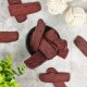 Eine schwarze Schüssel, gefüllt mit rechteckigen Hundeleckerlis, umgeben von verstreuten Leckerlis und zwei weißen Seilspielzeugen, auf einer grauen Oberfläche.