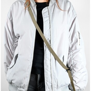 Eine Frau trägt eine hellgraue Bomberjacke über einem schwarzen Shirt, mit einem grünen Umhängegurt, vor einem schlichten Hintergrund.