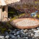 Ein Blaumeise schwebt über einer Holzschüssel mit Sonnenblumenkernen, neben einem Holzvogelhaus, umgeben von buntem Moos und Steinen.