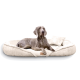 Ein grauer Weimaraner-Hund, der auf einem plüschigen, cremefarbenen Haustierbett mit einem kleinen passenden Kissen entspannt.