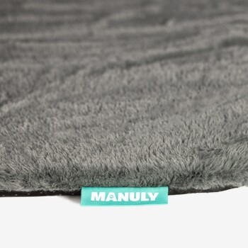 Rand einer grauen, plüschigen Haustiermatte mit türkisfarbener Stofflasche mit Aufschrift 'MANULY'.