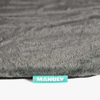 Rand einer grauen, plüschigen Haustiermatte mit türkisfarbener Stofflasche mit Aufschrift 'MANULY'.