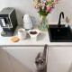 Eine graue Katze greift nach einer weißen Schüssel mit Haustierfutter auf einer Küchenarbeitsplatte, neben einer Kaffeemaschine, einem Wasserkocher und einer Vase mit Blumen.