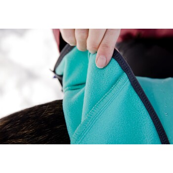 Eine Person hält eine türkisfarbene Haustierjacke, die ihren weichen Stoff und die Nähdetails zeigt, mit einem verschwommenen schneebedeckten Hintergrund.