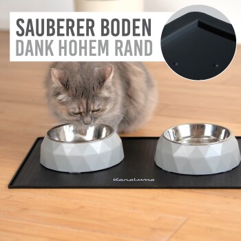 Eine graue Katze schnüffelt an einer geometrischen Futterschüssel für Haustiere auf einer schwarzen Matte, mit dem Text "SAUBERER BODEN DANK HOHEM RAND" darüber.
