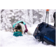 Ein Hund in einer türkisfarbenen und lila Jacke schnüffelt an einer blauen Ruffwear-Schüssel in einer verschneiten Landschaft, mit einer dunklen Patagonia-Tasche in der Nähe.