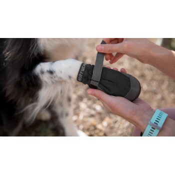 Eine Person zieht einem schwarzen Hundeschuh einen Hundepfote an, wobei der verstellbare Riemen und das 'Ruffwear'-Logo gezeigt werden.