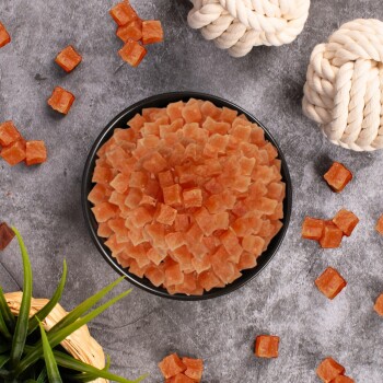 Eine schwarze Schüssel, gefüllt mit kleinen, orangefarbenen, würfelförmigen Haustierleckereien, umgeben von verstreuten Leckereien und zwei Seilspielzeugen auf einer grauen Oberfläche.