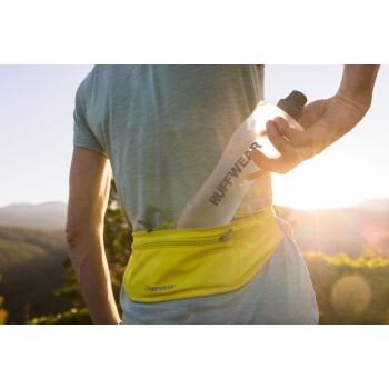 Eine Person in einem hellblauen Hemd und einer gelben Bauchtasche zieht eine klare Wasserflasche mit der Aufschrift "RUFFWEAR" aus der Tasche, mit einem Sonnenuntergang im Hintergrund.