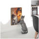 Eine verspielte graue Katze steht auf ihren Hinterbeinen und greift nach einem bunten, wandmontierten Kratzbrett mit einer Cartoon-Katze, die gegen einen Drachen kämpft.