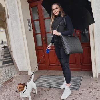 Eine Frau mit einem schwarzen Regenschirm führt einen kleinen braun-weißen Hund vor einem Gebäude aus, gekleidet in einer schwarzen Weste und Hose mit weißen Sneakers.
