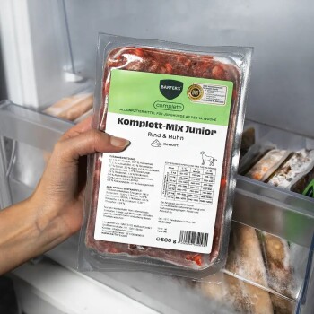 Hand hält vakuumversiegelte, gefrorene Futterverpackung 'BARFER'S complete Komplett-Mix Junior Rind & Huhn 500 g' im Gefrierschrank.