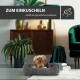 Ein kleiner Hund ruht sich in einem gemütlichen grauen Haustierbett aus, umgeben von stilvoller Wohnkultur, mit dem Text 'ZUM EINKUSCHELN schafft ein gemütliches Ambiente' darüber.